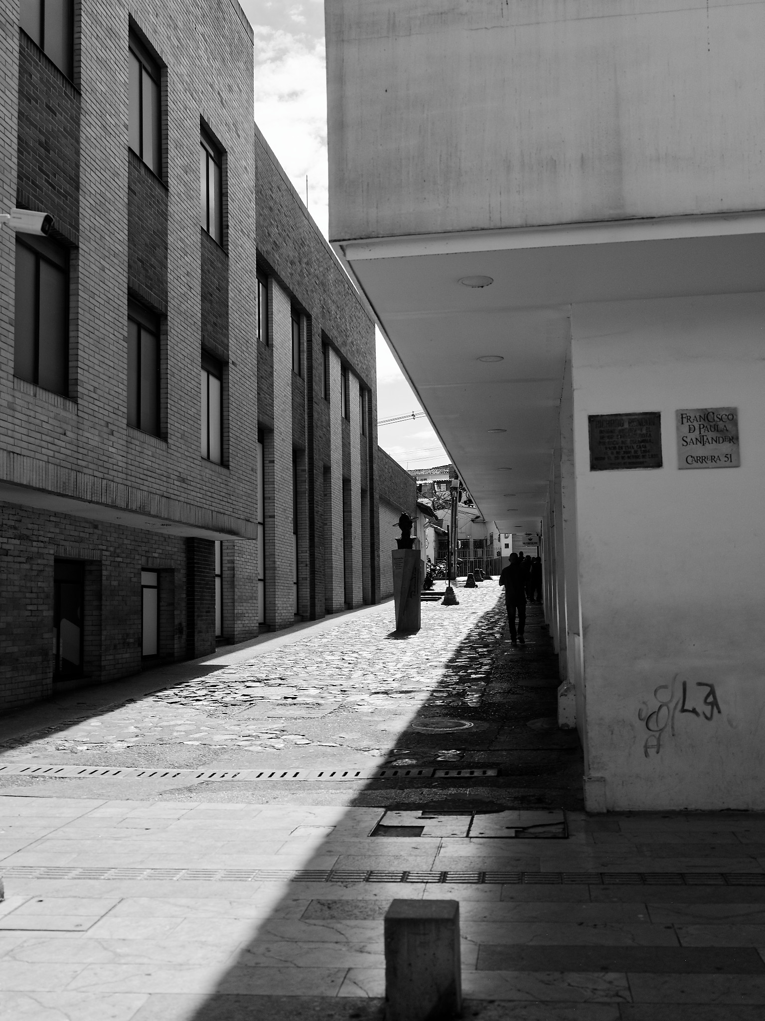 A narrow urban passage between brick and concrete buildings, divided by sunlight and deep shadow